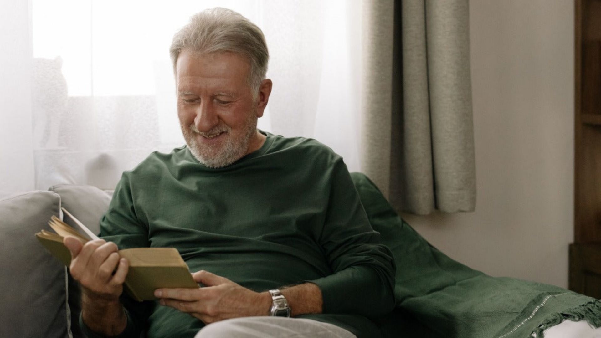 An elderly man on a couch, focused on reading a book about cannabis facts, surrounded by a cozy living room setting