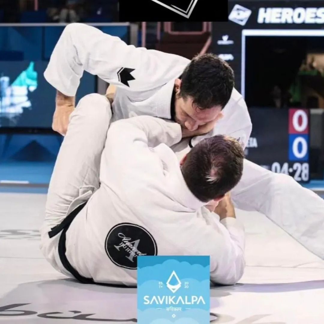 Two athletes in white gis grappling intensely on a mat during a Brazilian Jiu-Jitsu competition, with Savikalpa branding