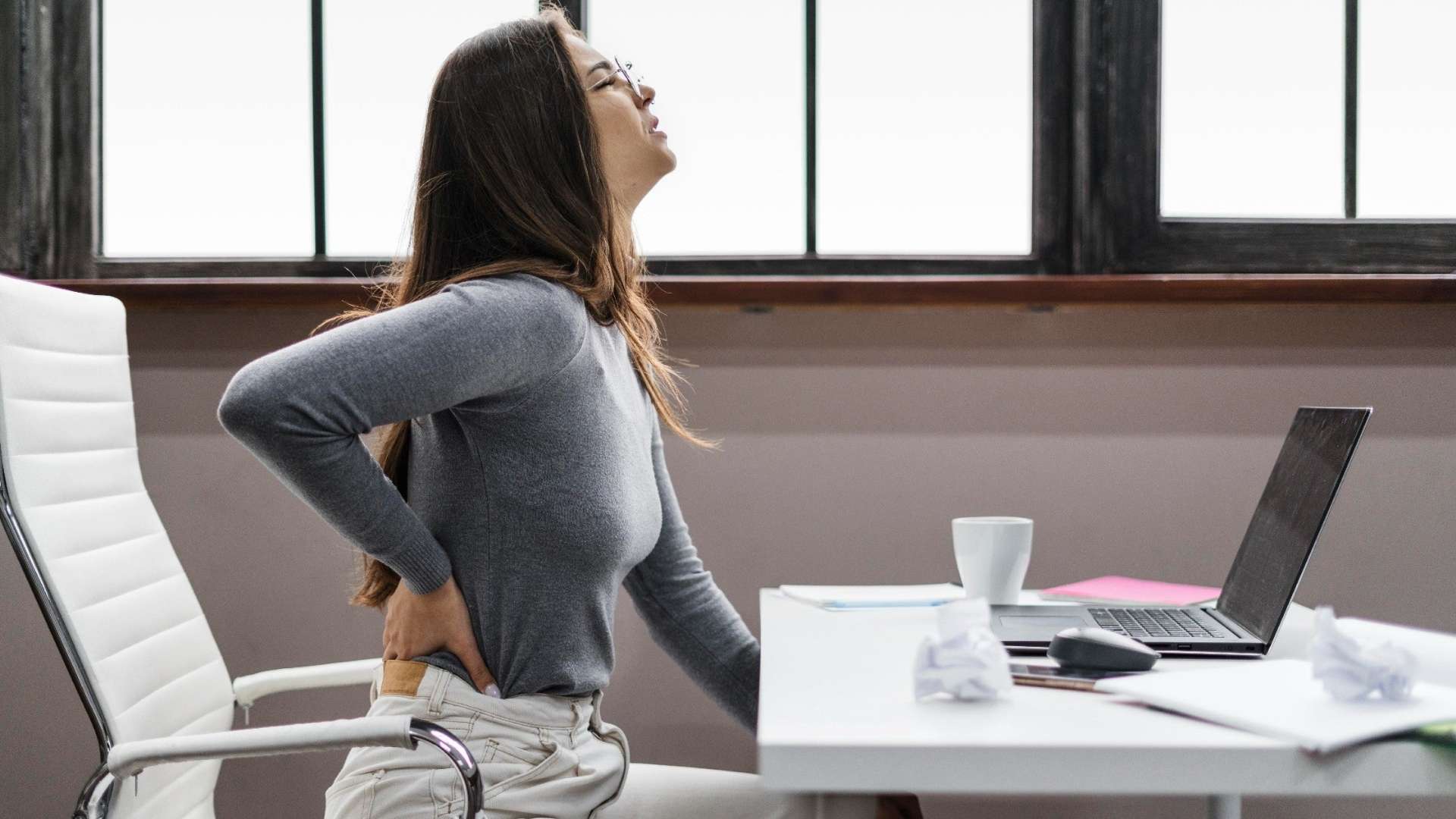 \An image of a young woman sitting on her work desk in front of her laptop and suffering from intense back pain