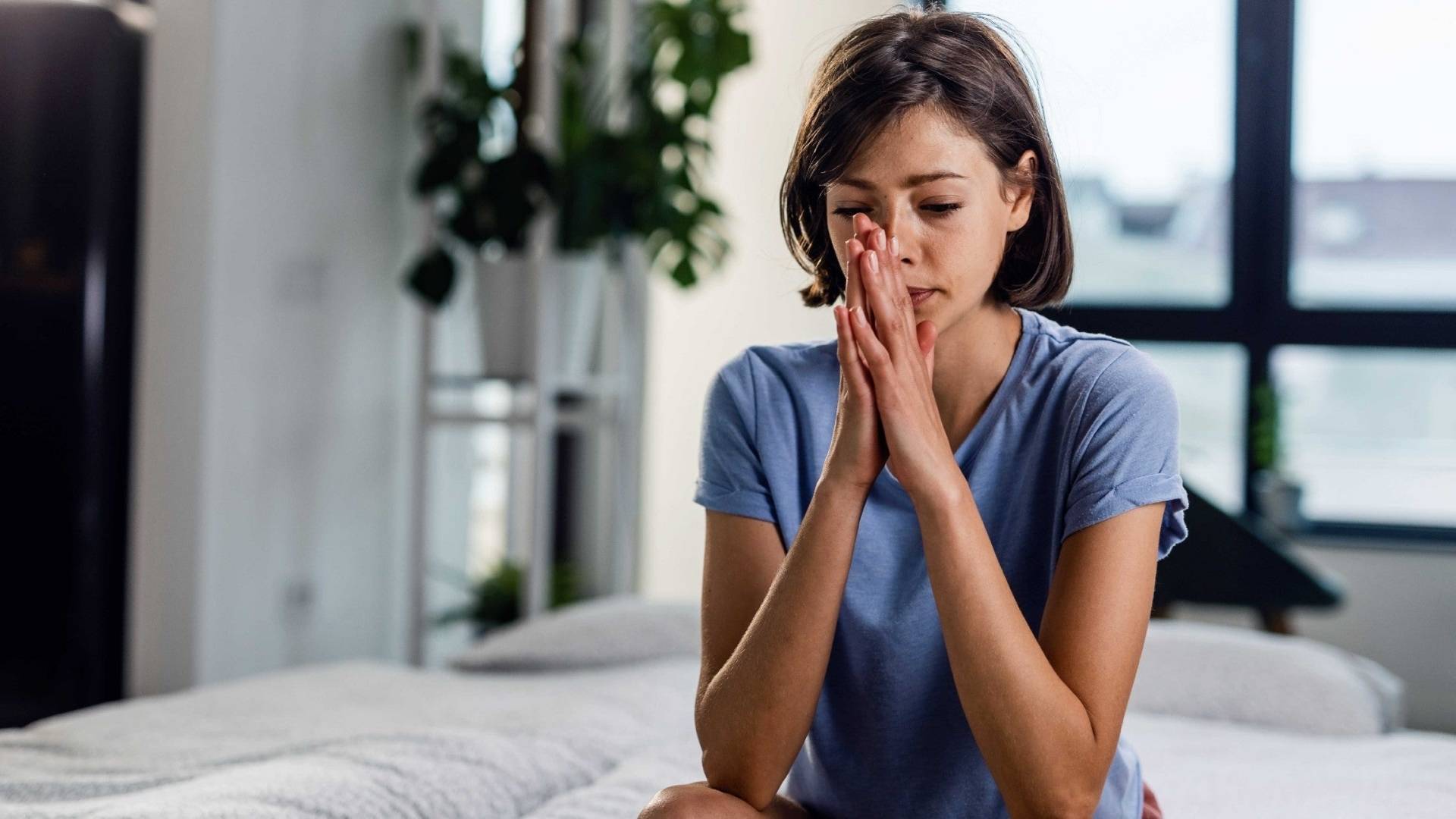 An image of a woman sitting on a bed, looking as,d with the backdrop of a bedroom setting and indoor plants