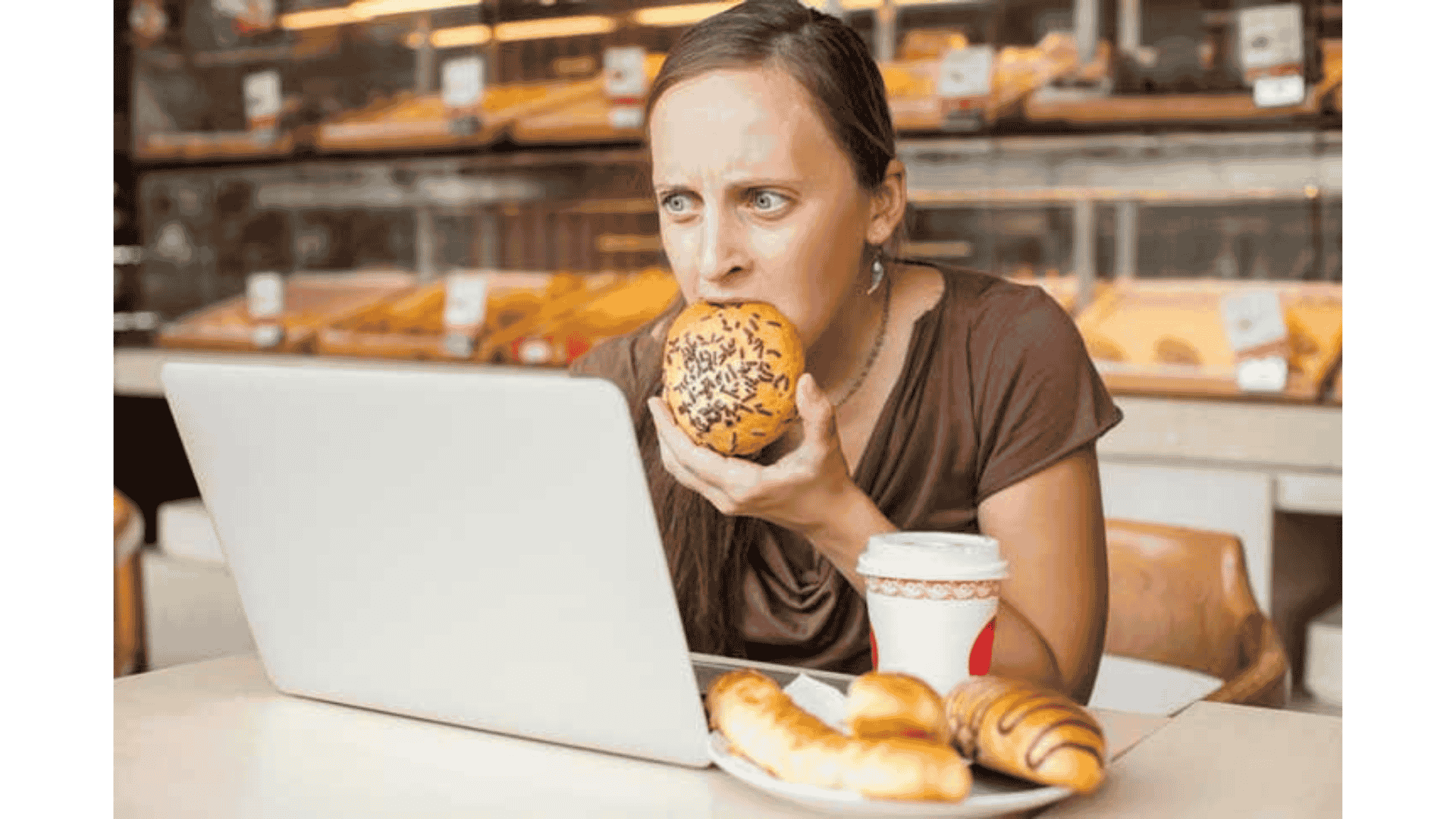 A woman sitting in a cafe in front of her laptop screen, eating a pain au chocolate ,showing appetite irregularity