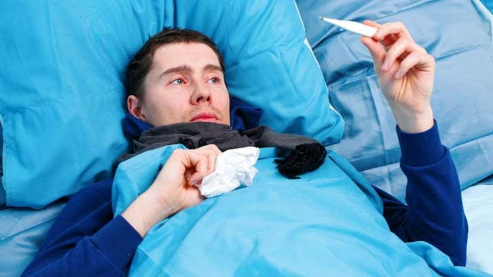 An image of a man lying in a bed checking his temperature with a thermometer, showcasing that he is sick