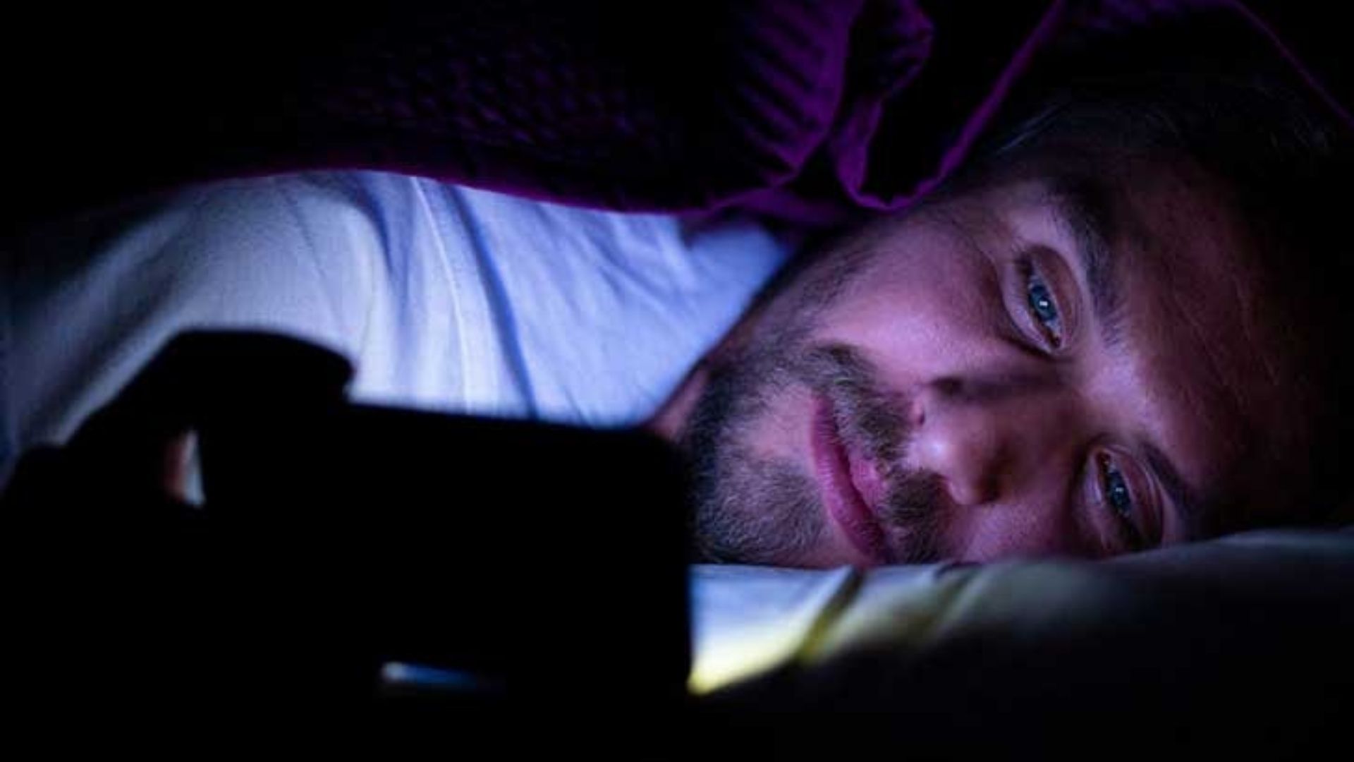 A man with insomnia lying in his bed at night with a mobile screen light being emitted, casting a blue colour