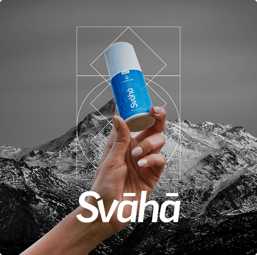 A hand elegantly holding a Svāhā topical pain cream bottle against a grey mountain background.